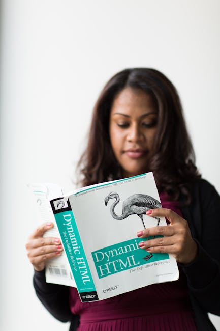 A woman with shoulder-length dark hair and light brown skin is standing indoors, holding an open book titled 'Dynamic HTML' by O'Reilly, which features an illustration of a flamingo on the cover. She 