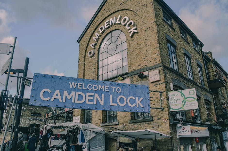 A street scene in Camden with a metal railway bridge overhead displaying a large painted sign reading 'Camden Lock' in bold yellow letters against a weathered blue background. The bridge features mura